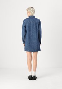 Jeansklänning i mörkblått, med långa ärmar, krage och sidofickor. Avslutad med knapppartier vid handlederna.