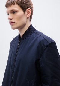 Μπουφάν βομβιστή σε ναυτικό μπλε με ριμπ γιακά, φερμουάρ μπροστά και ομοιόμορφη υφή. Διαθέτει κρυφές τσέπες και κλασική σιλουέτα.