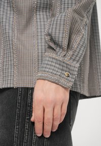 Camicia grigia a quadri con tessuto testurizzato, cuciture a contrasto e bottone sul polsino. La manica ha una vestibilità rilassata.