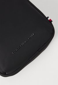 Svart laptopveske laget av et slitesterkt materiale, med en teksturert overflate og preget "TOMMY HILFIGER"-logo. Inkluderer en trikolor aksent.