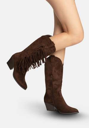 Piernas que llevan botas de vaquero de ante marrón oscuro hasta media pantorrilla, con detalles de flecos en una bota y tacones apilados en ambas botas.