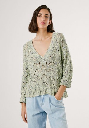 Lys grøn strikket sweater med V-hals, løs pasform, indviklet åbent mønster og rullede manchetter, parret med lyseblå jeans.