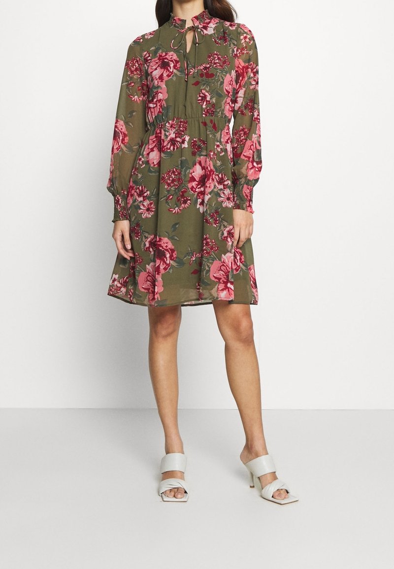 Femme portant une robe fleurie vert olive longueur genou avec manches longues et col à volants, associée à des sandales blanches à talons et ouvertes à l'avant.