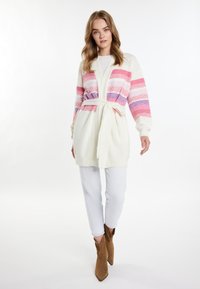 Strickjacke mit pink-und lila gestreiftem Muster, cremefarbener Basis, Knotenbund am Saum, lange Ärmel, kombiniert mit weißen Hosen und braunen Stiefeletten.