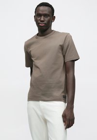 Καφέ βαμβακερό μπλουζάκι με κοντά μανίκια, στρογγυλή λαιμόκοψη και μικρή ετικέτα στο τελείωμα· σε συνδυασμό με λευκό παντελόνι.