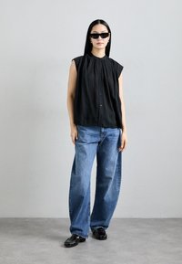 Blouse noire sans manches avec col froncé, associée à un jean large bleu clair et des mocassins noirs. Comprend des lunettes de soleil.