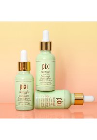 Tre glassflasker med PIXI Overnight Glow Serum i lys grønn, med gull dråpetopper og etiketter, plassert på en gradient bakgrunn.