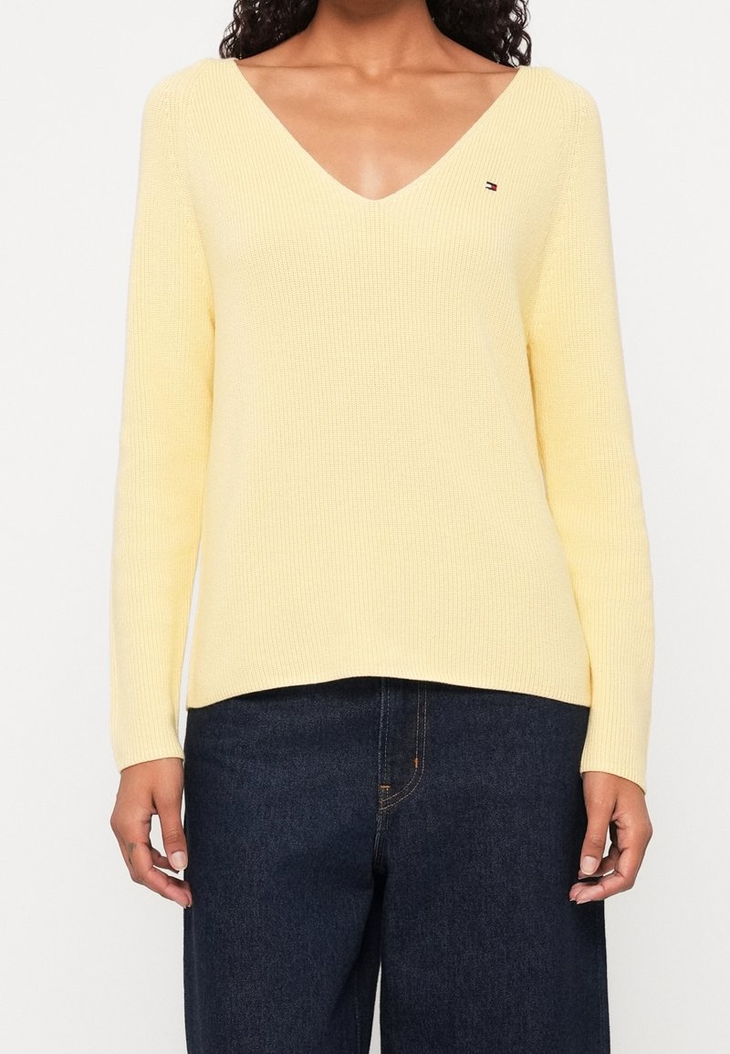 Femme portant un pull en maille jaune clair à col en V avec manches longues et un jean bleu foncé, debout devant un fond blanc.