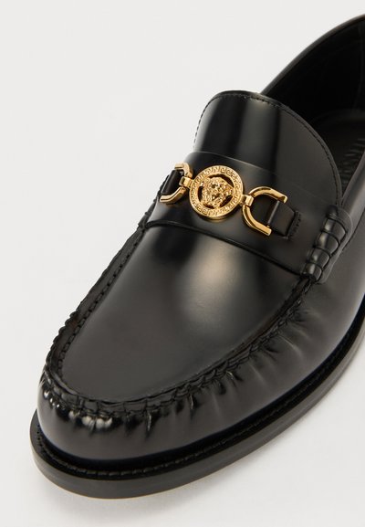 Svarta läder loafers med en guldmedaljong med lejonmönster och dekorativ sömnad längs sidorna. Slät yta och rundad tå.