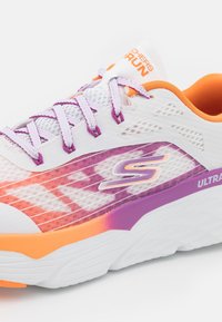 Zapatilla deportiva blanca con acentos en púrpura y naranja, que presenta una parte superior de malla, un patrón texturizado y una suela acolchada. Logo notable en el lateral.