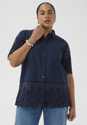 Žena v tmavomodrej košeli na gombíky s kvetinovým čipkovaným rukávom a lemom, v svetlomodrých džínsoch, pózujúca s rukou pri krku.