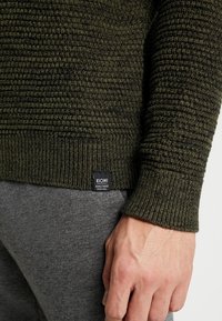 Manche et ourlet d'un pull en maille vert foncé avec une étiquette "KIOMI", assortis à un pantalon gris et une main détendue visible.
