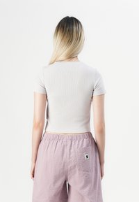 Pikkade blonde juustega inimene, kellel on seljas beež lühikeste käistega topp ja heledad lilla tekstuuriga lühikesed püksid, millel on tagatasku ja logo silt.