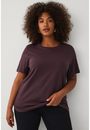 Vrouw met krullend haar die een donkerpaars kort mouw shirt en zwarte broek draagt, staand voor een effen lichte achtergrond.