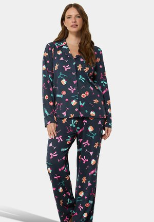 Femme aux longs cheveux bruns portant un pyjama bleu marine foncé décoré de bonbons de Noël colorés, de cannes en sucre d'orge, de bonshommes en pain d'épices et de rubans.