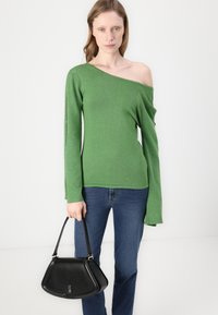 Grønn, off-shoulder genser med lange ermer, laget av et teksturert stoff. Svart håndveske med en strukturert form, holdt i høyre hånd.
