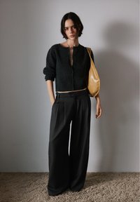 Jonge vrouw in een zwarte cropped cardigan en zwarte broek met wijde pijpen, met een tan schoudertas, staand op een tapijtvloer tegen een effen muur.