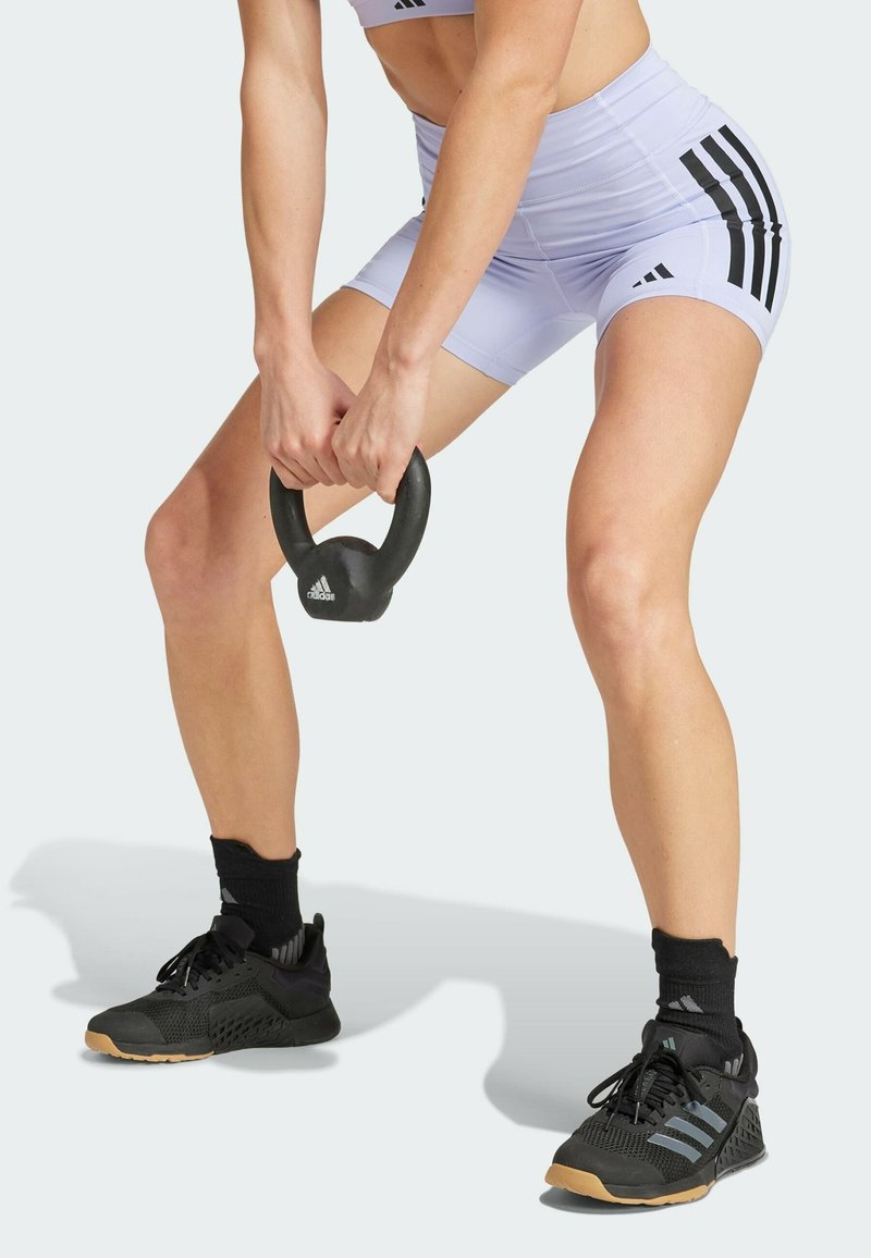 Mustaks kettlebell, mida hoiab kahe käega, seljas on heledat lilla värvi lühikesed püksid ja mustad spordijalatsid. Pükstel on külgedel mustad vertikaalsed triibud.