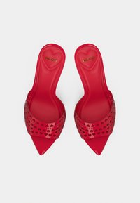 Sandales rouges à enfiler avec un bout pointu, présentant un motif de cœur perforé sur la bride et une finition brillante.