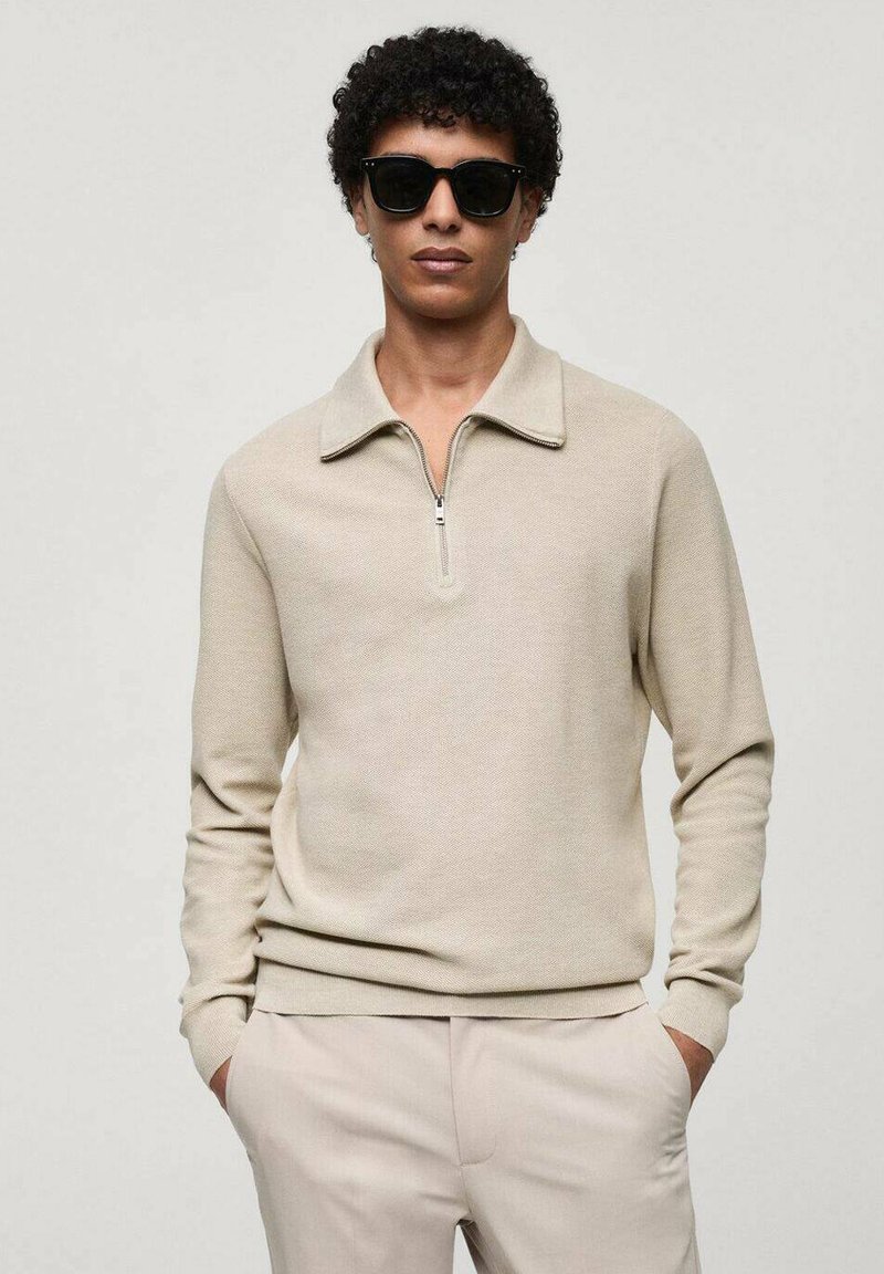 Beige langærmet pullover med lynlås i kraven, lavet af tekstureret stof. Båret med lyse bukser og mørke solbriller.