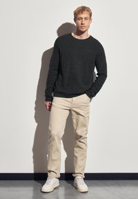Homme adossé à un mur blanc, portant un pull noir, un pantalon beige et des baskets blanches, avec une main dans la poche.