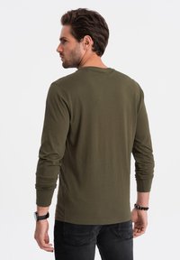 Langärmliges olivgrünes T-Shirt aus weicher Baumwolle. Mit Rundhalsausschnitt und schmalen Ärmeln. Getragen zu schwarzen Jeans und einem Armband.