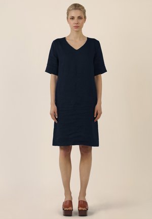 Vrouw staand, naar voren kijkend, gekleed in een knie-lange marineblauwe V-hals jurk en bruine platform open-toe schoenen tegen een effen achtergrond.