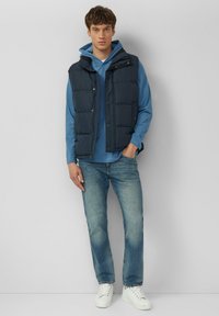 Granatowa kamizelka puchowa na jasnoniebieskiej bluzie z kapturem, w połączeniu z wyblakłymi niebieskimi dżinsami i białymi trampkami; posiada zatrzaski i usztywnioną konstrukcję.