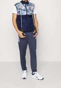 Námořnická prošívaná vesta s vlnkovým vzorem, v kombinaci s námořnickými tapered joggers. Obsahuje bílé tenisky a odporové gumy v ruce.
