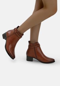 Piernas de mujer usando botas de cuero marrón hasta el tobillo, con cremallera lateral y correas decorativas, sobre un fondo blanco.