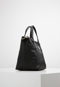 Sort tote taske med dobbelt læderhåndtag, guldfarvet hardware og et rundt logoemblem foran, præsenteret på en hvid overflade.