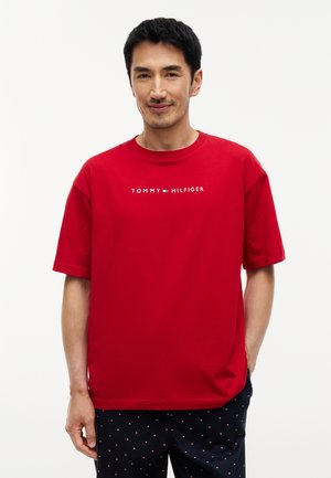 Man draagt een rood Tommy Hilfiger T-shirt en een zwarte broek met kleine witte en rode patronen, staand tegen een effen achtergrond.