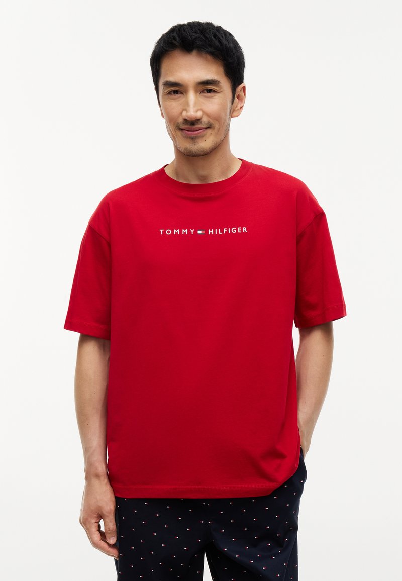 Homme portant un t-shirt rouge Tommy Hilfiger et un pantalon noir avec de petits motifs blancs et rouges, debout devant un fond uni.