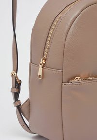 Sac à dos en cuir beige avec une forme arrondie, une fermeture éclair dorée, une sangle réglable et une petite poche avant. Surface à texture lisse.