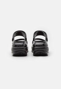 Zuecos de plástico negro y brillante con correas traseras ajustables y un diseño perforado para ventilación. Suelas texturizadas para un mejor agarre.