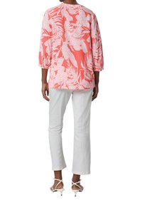 Koralbluse mit einem weißen Blumenmuster, dreiviertel Ärmel; kombiniert mit weißen, engen Hosen und weißen, schnürbaren Heels. Glatte Stofftextur.
