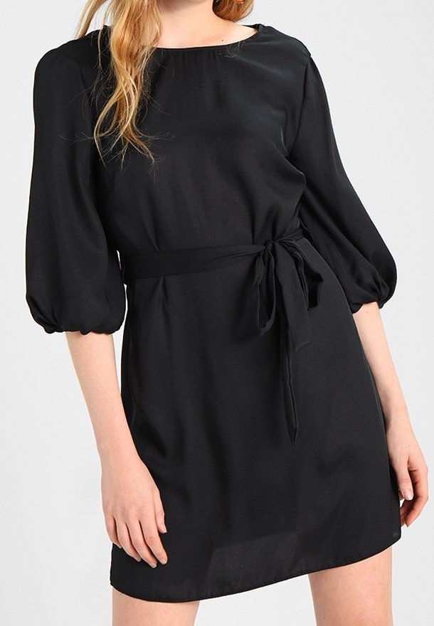 Femme portant une robe noire longueur genou avec des manches bouffantes trois-quarts et une ceinture nouée à la taille sur un fond uni.