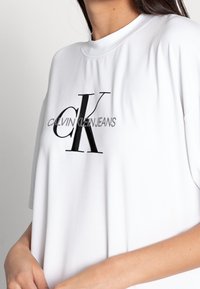 Woman wearing white Calvin Klein Jeans t-shirt with large black "cK" logo on chest, holding the bottom of the shirt.