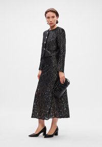 Juoda nėriniuota suknelė su prigludusia liemeniu ir ilgomis rankovėmis, puošta metaliniais siūlų raštais; derinama su juodu klostuotu rankiniu maišu ir bateliais.