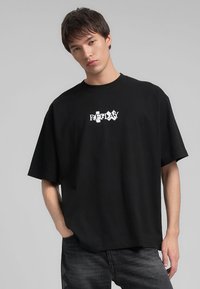 T-shirt oversize in cotone nero con maniche corte, caratterizzata da un logo grafico bianco sul petto. Tessuto morbido, vestibilità casual.