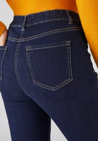 Jeans de denim azul oscuro con un diseño ajustado, que cuentan con una cinturilla elástica alta y costuras doradas en contraste en los bolsillos.
