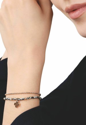 Zwei Perlenarmbänder an einem Handgelenk, eines mit einem vierblättrigen Kleeblatt-Anhänger, vor dem Kinn einer Person und schwarzem Ärmelhintergrund.