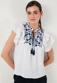 Balta bluzelė su tamsiai mėlynu gėlių siuvinėjimu ir kutais. Yra raukiniai ant rankovių ir kaklo spragų dizainas. Lengvas audinio tekstūro.