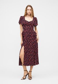 Γυναίκα που φοράει μια μπλε midi φόρεμα με κόκκινες ανθισμένες εκτυπώσεις, με φουσκωτά μανίκια, πλαϊνές σκισμές και μαύρα σανδάλια με ψηλά τακούνια και λουράκια, στέκεται μπροστά σε απλό φόντο.