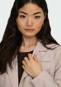 Lekka beżowa marynarka ze sztruksu o ustrukturyzowanym kształcie i subtelnych detalach szycia, noszona na ciemnobrązowym topie. Widoczna miękka, teksturowana tkanina.