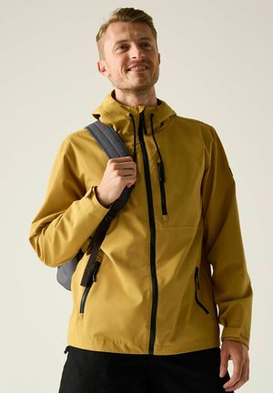 Glimlachende man met een mosterdgele capuchontrui en zwarte broek, met een grijze rugzak op één schouder tegen een neutrale achtergrond.