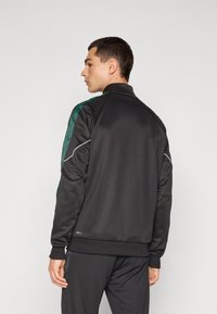 Veste de sport noire avec des accents verts à motifs sur les épaules, texture lisse, col montant et passepoil blanc le long des épaules ; portée avec un pantalon assorti.