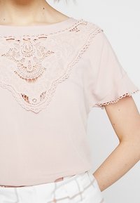 Ljusrosa blus med korta ärmar, med en detaljerad spetspanel vid halsringningen och scalloped spetskant. Mjukt tyg och lös passform.