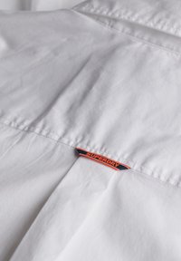 Camisa de algodón blanca con una textura suave. Presenta una etiqueta con "SUPERDRY" en naranja y azul marino, cosida en la parte posterior del cuello.