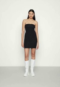 Mulher com longos cabelos escuros vestindo um vestido mini preto sem alças e botas brancas até ao joelho de sola grossa, em pé contra um fundo claro e liso.